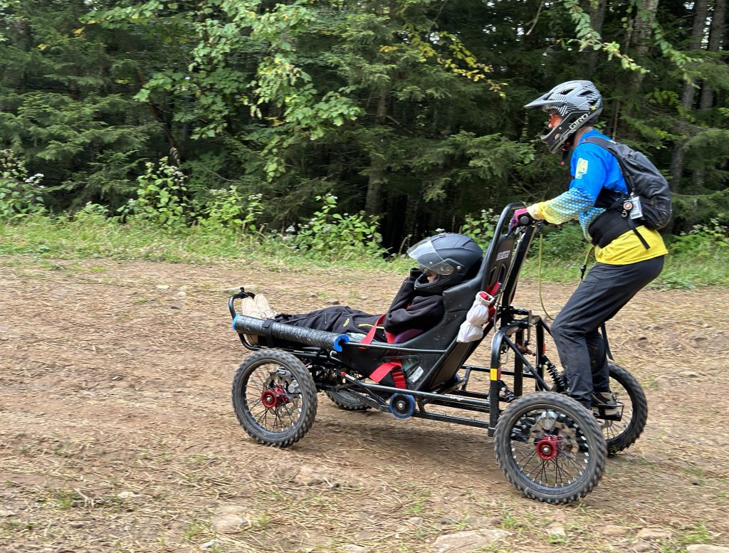 Carol Woody steers the Cimgo while standing firmly on the foot pegs at the back of the all-terrain device. Gage Tatar rides in the Cimgo giggling and smiling his way down the dirt path at Snowshoe Resort.