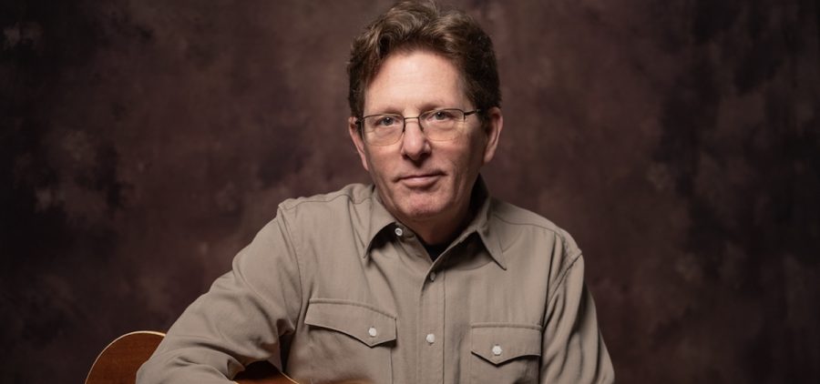 A promotional image of musician Tim O'Brien. He is posed with his guitar against a brown background.