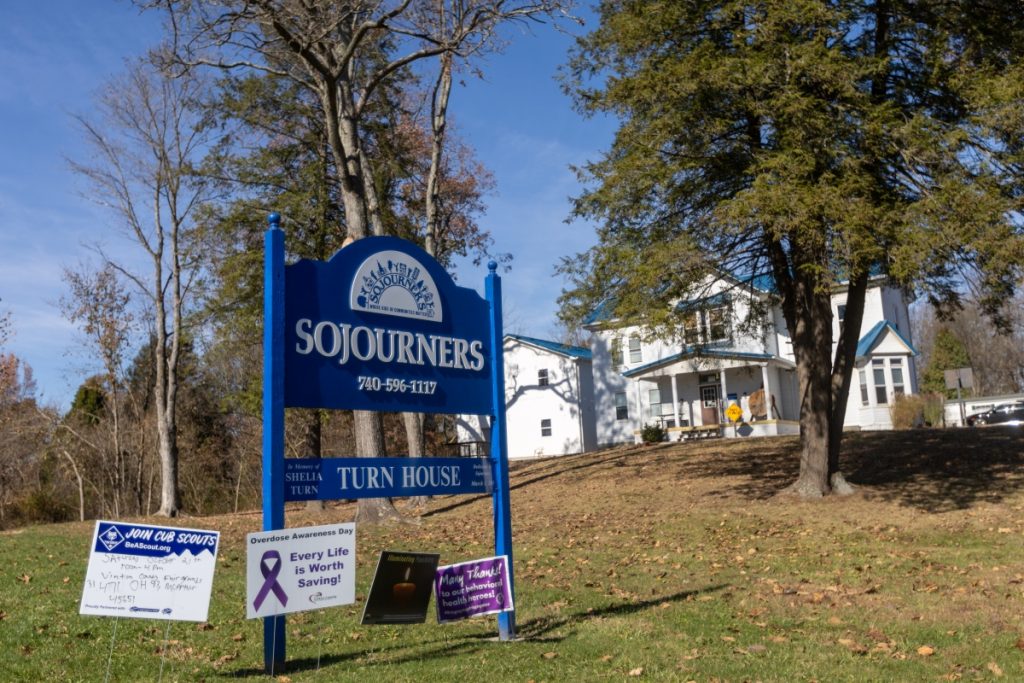 A large blue sign shares the phone number for Sojourners Care Network. There are two large houses in the background.