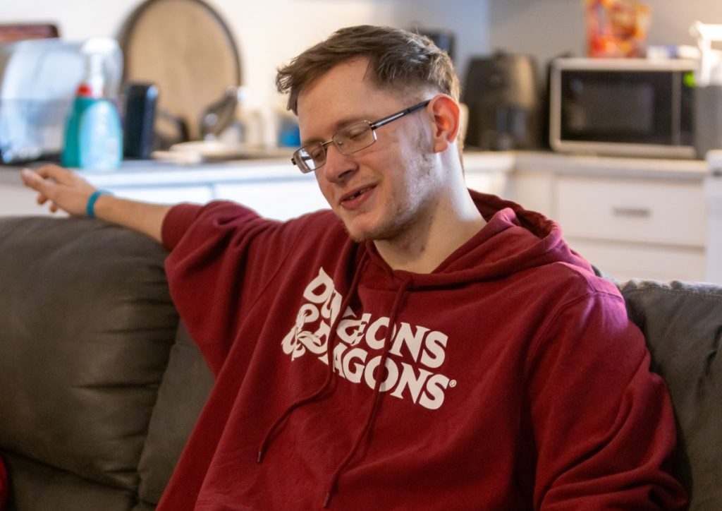A young man in a Dungeons & Dragons hoodie sits on a couch, smiling.