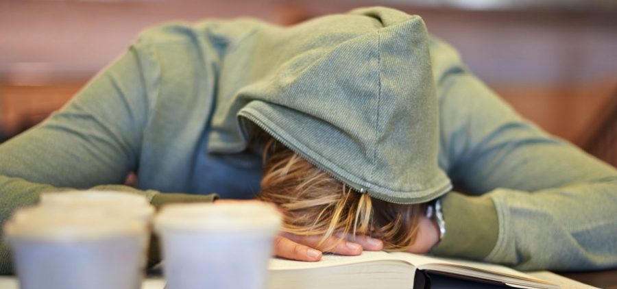 A young person wearing a hoodie over their head lays their head down on an open book.