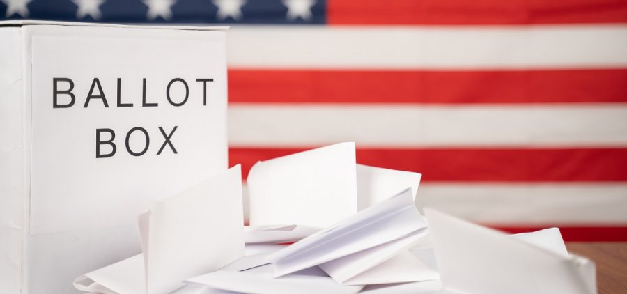 A ballot box has election results coming out of it with an american flag in the background.