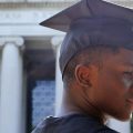 Close up of black MIT graduate in cap and gown with university building in background