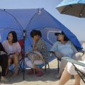A group of expectant mothers sit on the beach under an umbrella.