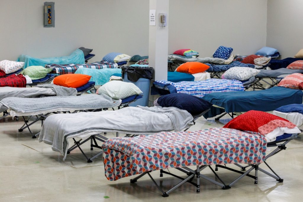 Rows of cots with blankets and pillows in a large open facility.