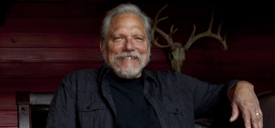 A promotional image of Jorma Kaukonen. he is standing up and wearing a sweater and a blazer.