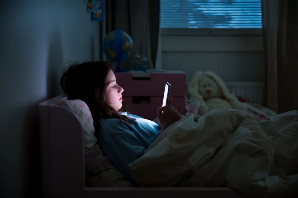 A girl lies in bed with the lights out, looking at her phone.