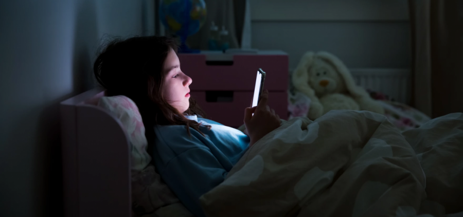 A girl lies in bed with the lights out, looking at her phone.