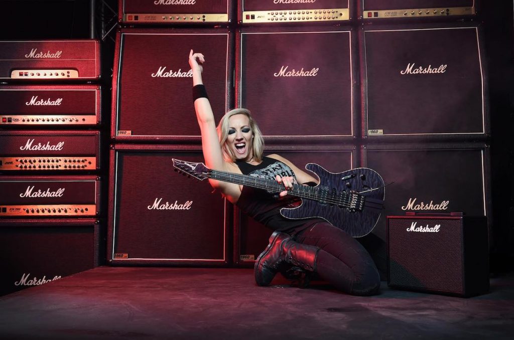 A press image of image of guitarist Nita Strauss posing in front of stacks of amps with a guitar in her hand. 