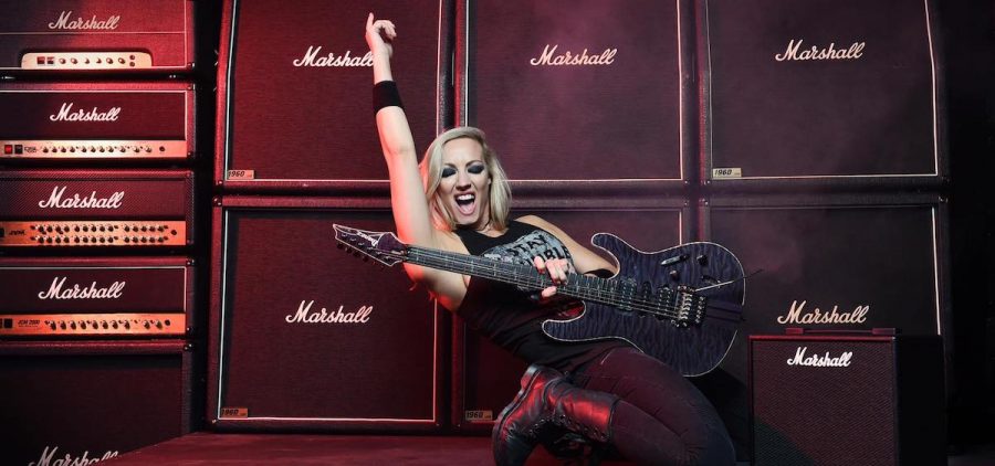 A press image of image of guitarist Nita Strauss posing in front of stacks of amps with a guitar in her hand.