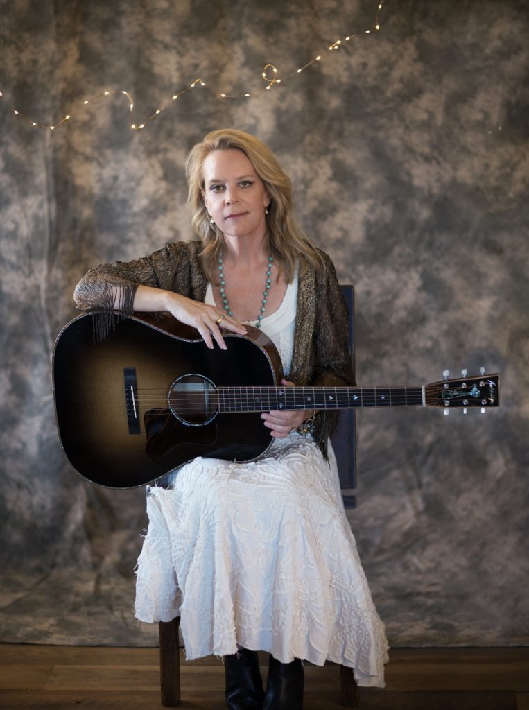 A promotional image of musician Mary Chapin Carpenter. She is sitting down and holding a guitar. 
