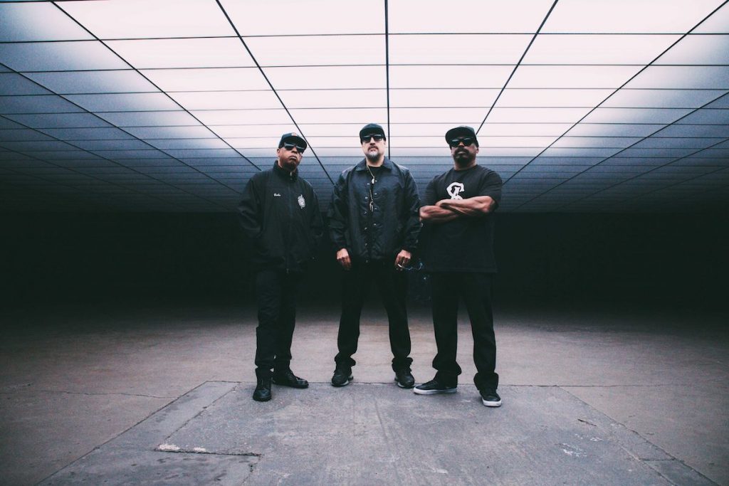 A promotional image of the band Cypress Hill. All three members are standing in a dark room. 