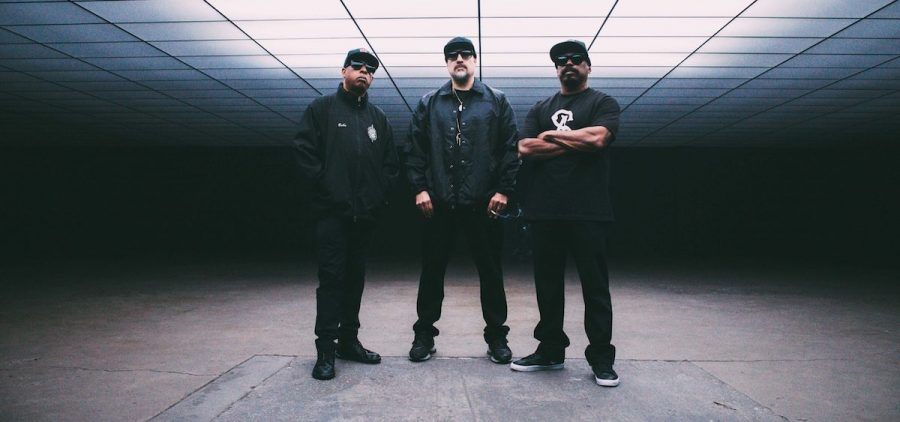 A promotional image of the band Cypress Hill. All three members are standing in a dark room.