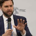 Ohio Senator JD Vance holding a microphone and his left hand up as he speaks at an international security conference in Germany.