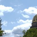 The West Virginia Capitol dome is seen through some trees with a blue sky and a few clouds behind.