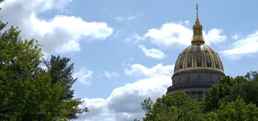 The West Virginia Capitol dome is seen through some trees with a blue sky and a few clouds behind.