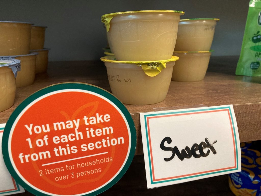 Cups of applesauce sit on a shelf. A sticker reads "You may take 1 of each item from this section."