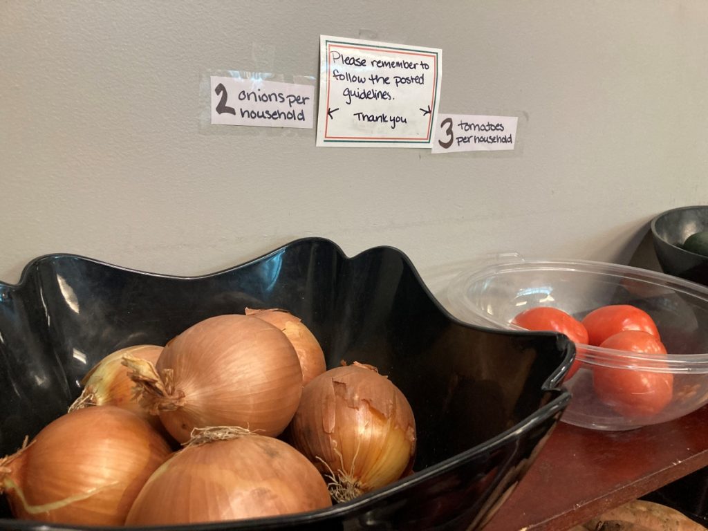 Onions sit in a black plastic bowl. Signs above the bowl read "2 onions per household" and "Please remember to follow the posted guidelines."