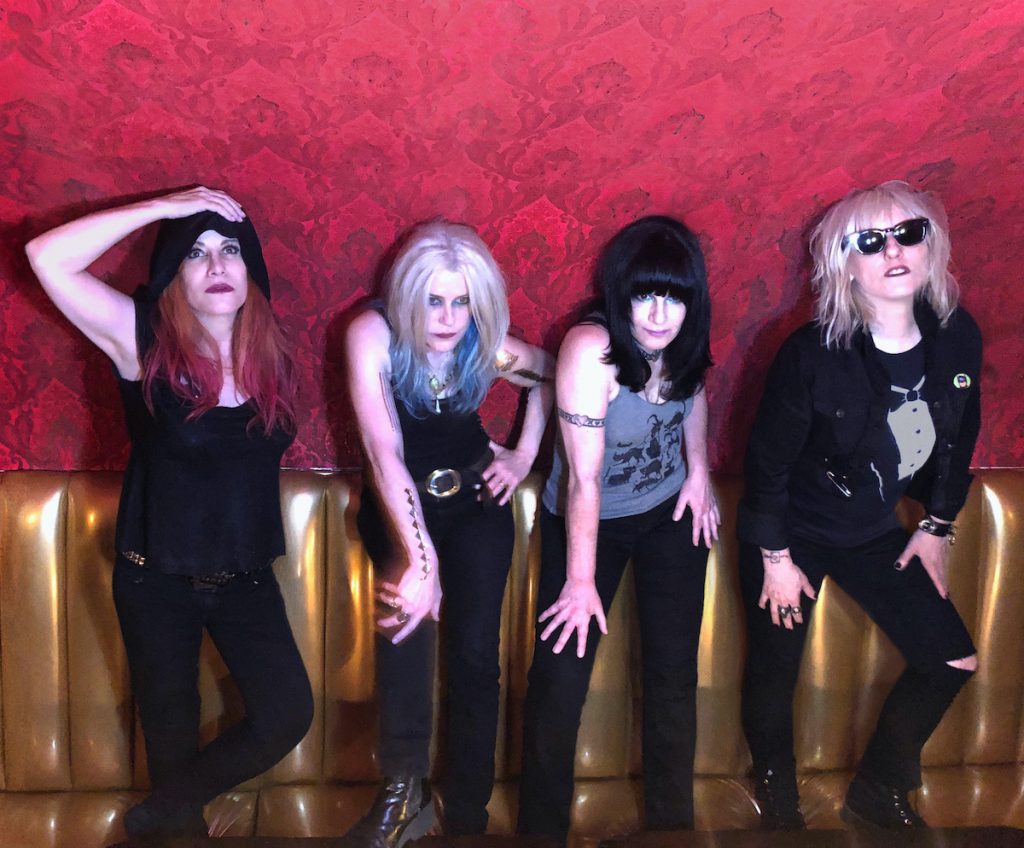 A promotional photo of the band L7. All four members are leaning against a red wall. 