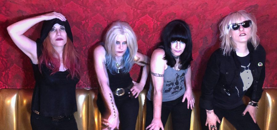 A promotional photo of the band L7. All four members are leaning against a red wall.