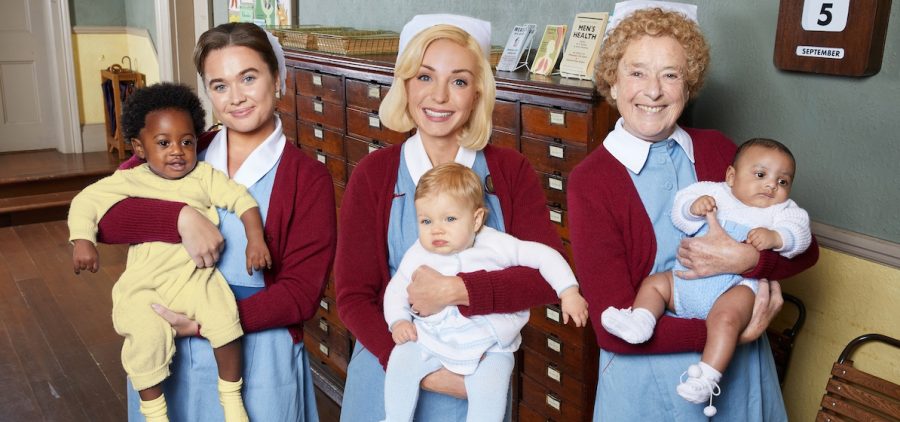 Call The Midwife S13. Three nurse midwives in uniform with red sweaters and blue dresses, each holding a baby.