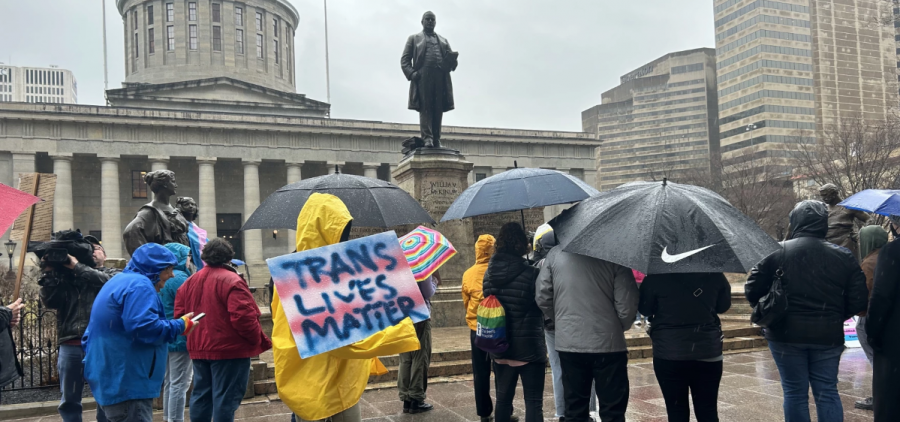 A group of people stand outside the Ohio Statehouse. One person holds a sign reading "Trans Lives Matter."
