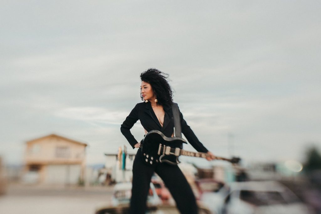 A promotional image of musician Judith Hill. She is standing against a desert landscape, dressed in black, with a guitar. 