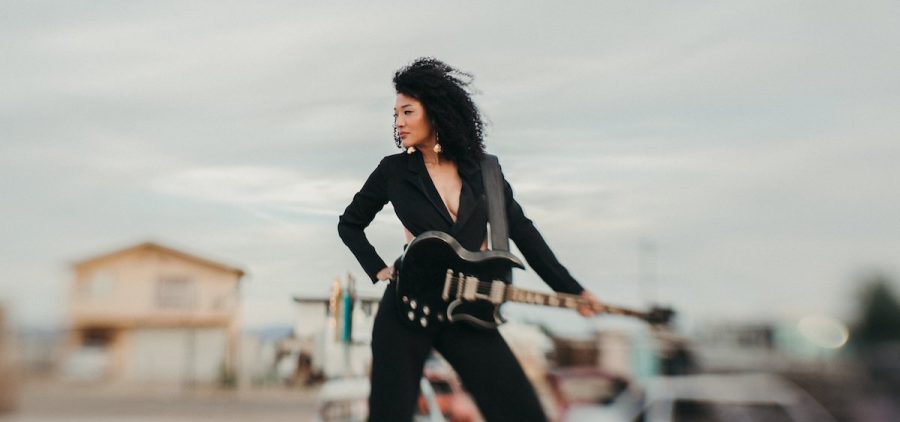 A promotional image of musician Judith Hill. She is standing against a desert landscape, dressed in black, with a guitar.
