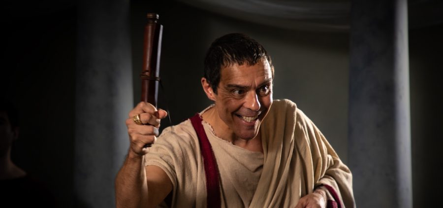 Caesar (Andonis Anthony) holds scroll with evil/happy smile on his face. Dark senate background Credit: Richard Pearson; BBC Studios