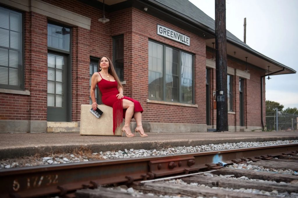 A promotional image of Eden Brent. She is sitting at a train station by herself in a red dress. 
