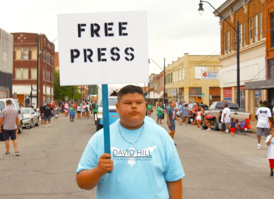 A film still from the film "Bad Press" showing a boy holding a sign reading "Free Press."