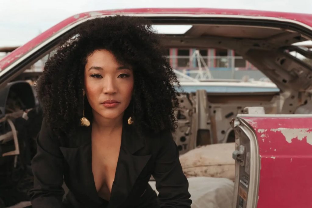 A promotional image of musician Judith Hill, sitting in a car wearing a black suit. 