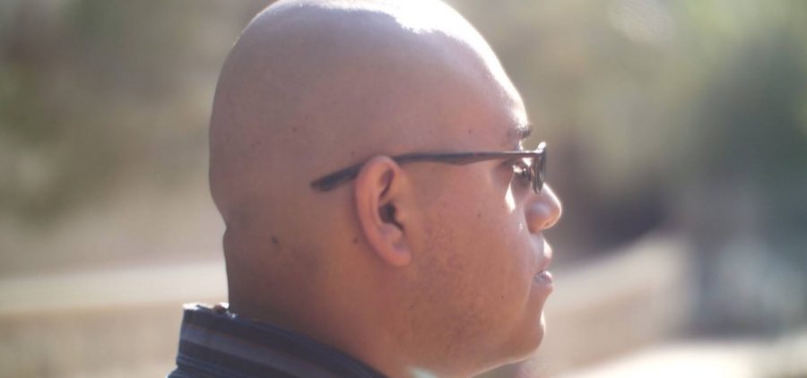 Side profile shot of Pedro, a bald young man with glasses wearing a black collared shirt on a sunny day. Credit: Set Hernandez