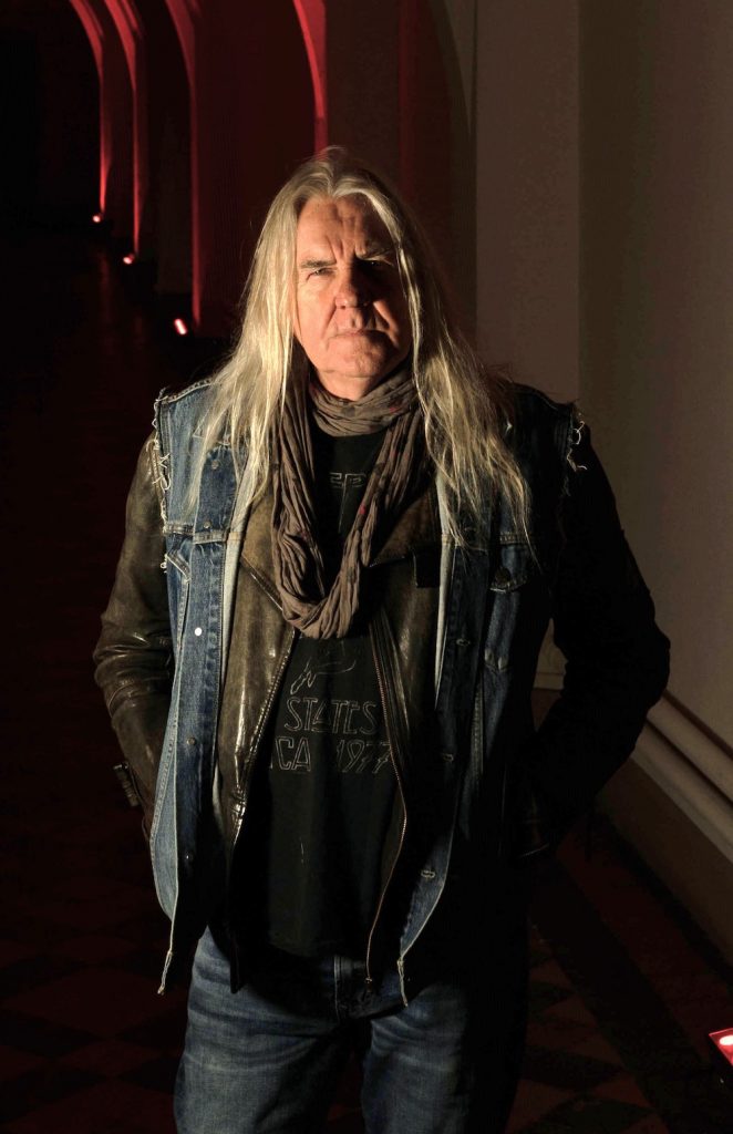 A promotional image of Biff Byford, the lead singer of Saxon. He is wearing black and is posed against a wall. 