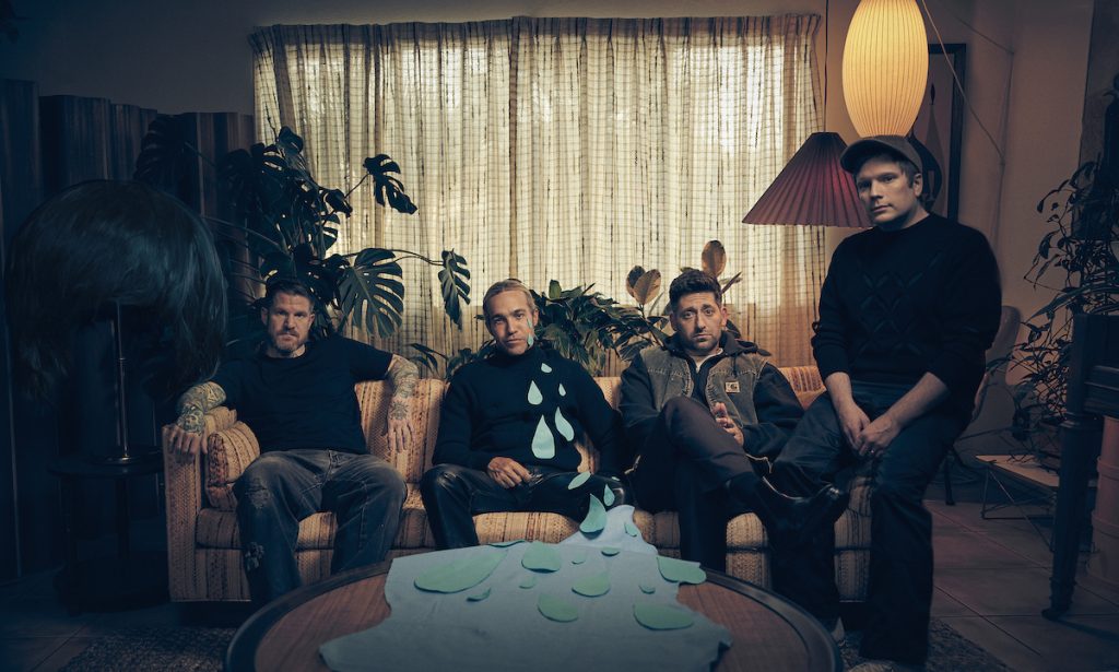 A promotional image of the band Fall Out Boy, all seated on a couch in a dark room. 