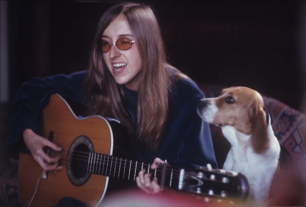 A woman with long hair and glasses plays her guitar by a beagle. 