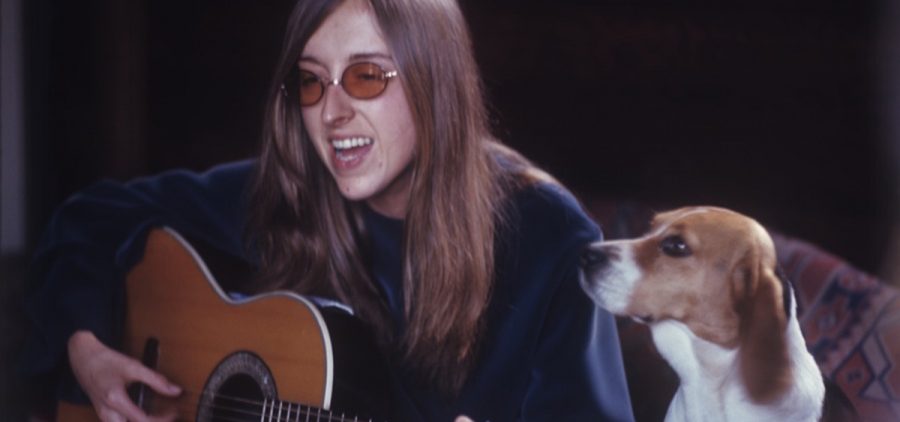 A woman with long hair and glasses plays her guitar by a beagle.