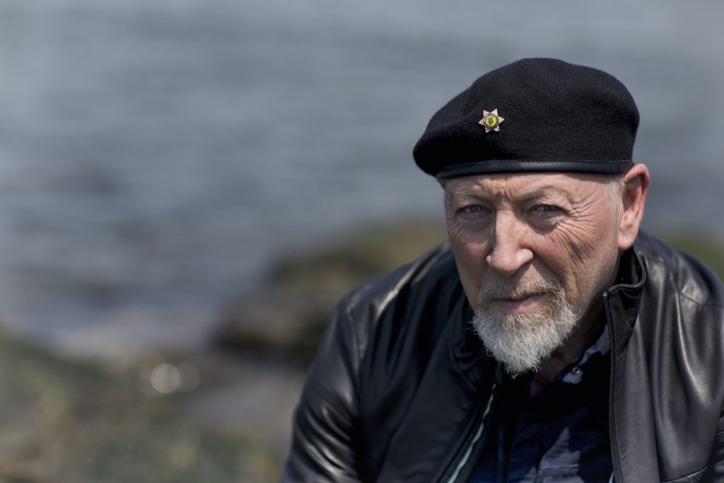 A promotional image of musician Richard Thompson. He is sitting outside by the sea. 
