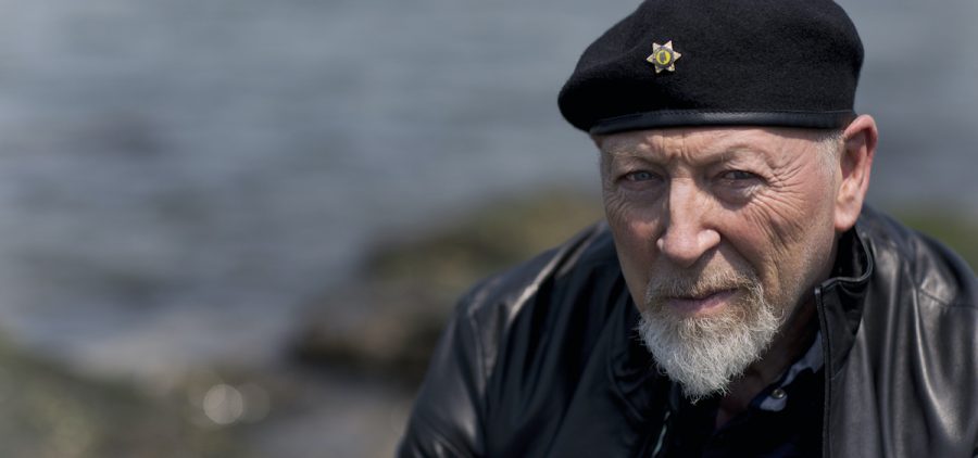 A promotional image of musician Richard Thompson. He is sitting outside by the sea.