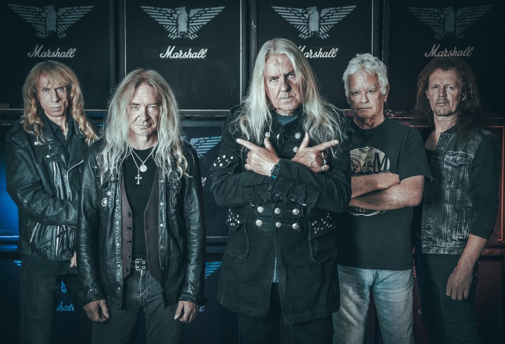 A promotional image of the metal band Saxon. They are all wearing black and are posed against large amplifiers. 