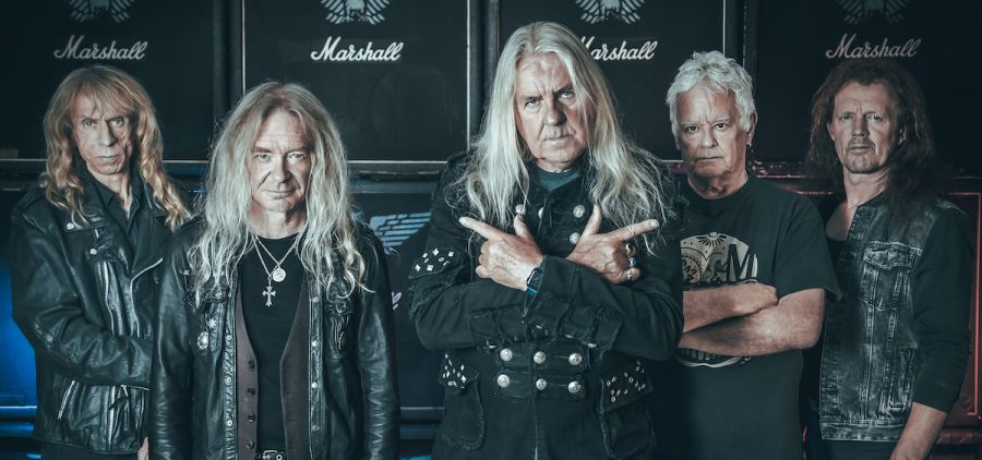 A promotional image of the metal band Saxon. They are all wearing black and are posed against large amplifiers.