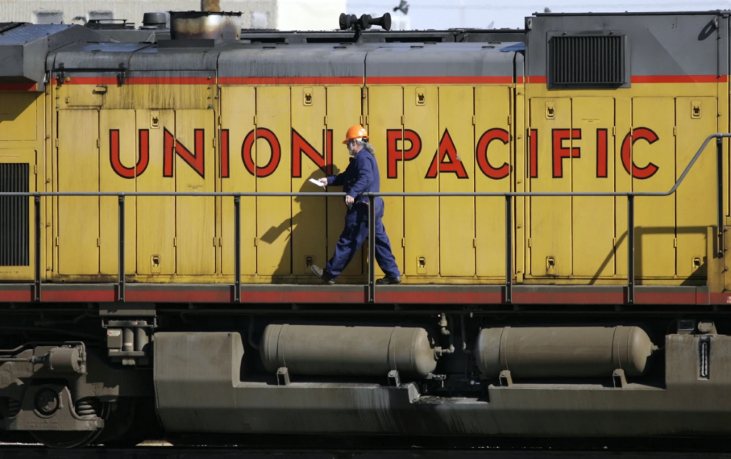 A man in a blue uniform and orange safety helmet walks in front of a yellow Union Pacific train car.