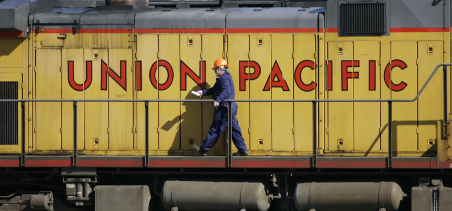A man in a blue uniform and orange safety helmet walks in front of a yellow Union Pacific train car.