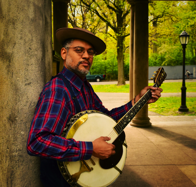 A promotional image of musician Dom Flemmins. He is outside and holding a banjo under a tree.