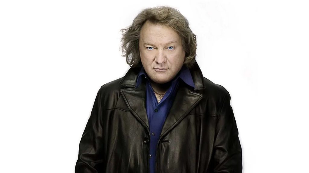 A promotional image of former Foreigner singer Lou Gramm. He is against a white background and wearing a leather jacket. 