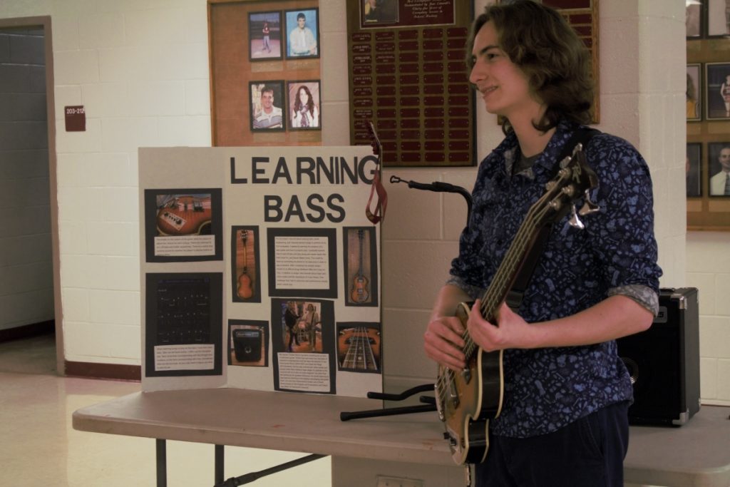A high school student stands next to a poster, ready to play a bass guitar.