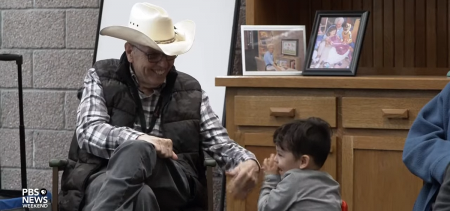 A smiling older man in a cowboy hat reaches his hand out to a child at a meeting of kids and seniors.