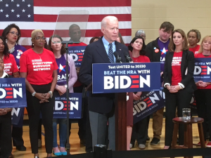 President Joe Biden stands at a podium that reads "BEAT THE NRA WITH BIDEN." Several people hold Biden signs behind him.