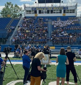 A crowd gathers at a high school athletics field to watch a press conference.