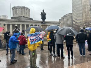Ohioans gather in the rain outside the Ohio Statehouse while the Ohio Senate overrides Gov. Mike DeWine's veto of a bill that limits health care for trans kids and bans trans kids from competing on girls sports teams.